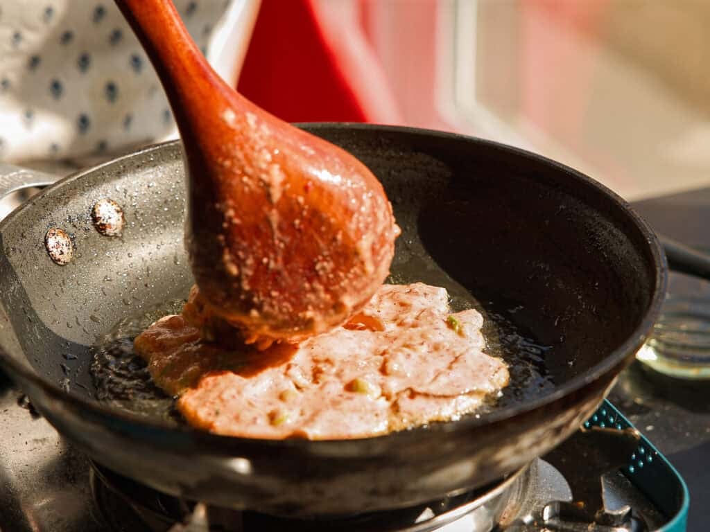 A person uses a wooden spoon to spread kimchi pancake mixture in a frying pan on a stovetop, with sunlight illuminating the scene.